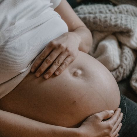 Schwangere Frau auf dem Sofa sitzend, hält ihren runden Babybauch und blick glücklich auf ihre Hände