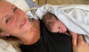 Glückliche Mutter liegt im Krankenhausbett und lächelt in die Kamera, hat ihr Neugeborenes auf der Brust liegen